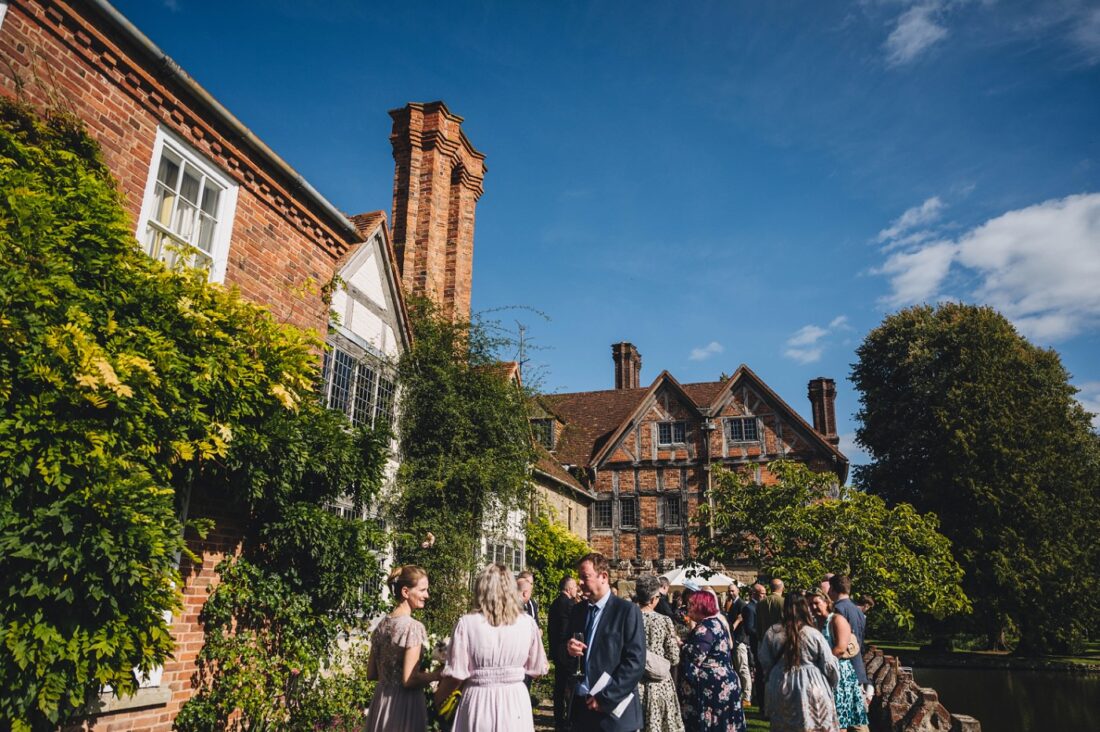 wedding guests enjoying drinks on the terrace at Birtsmorton court by the moat, blue skies above them