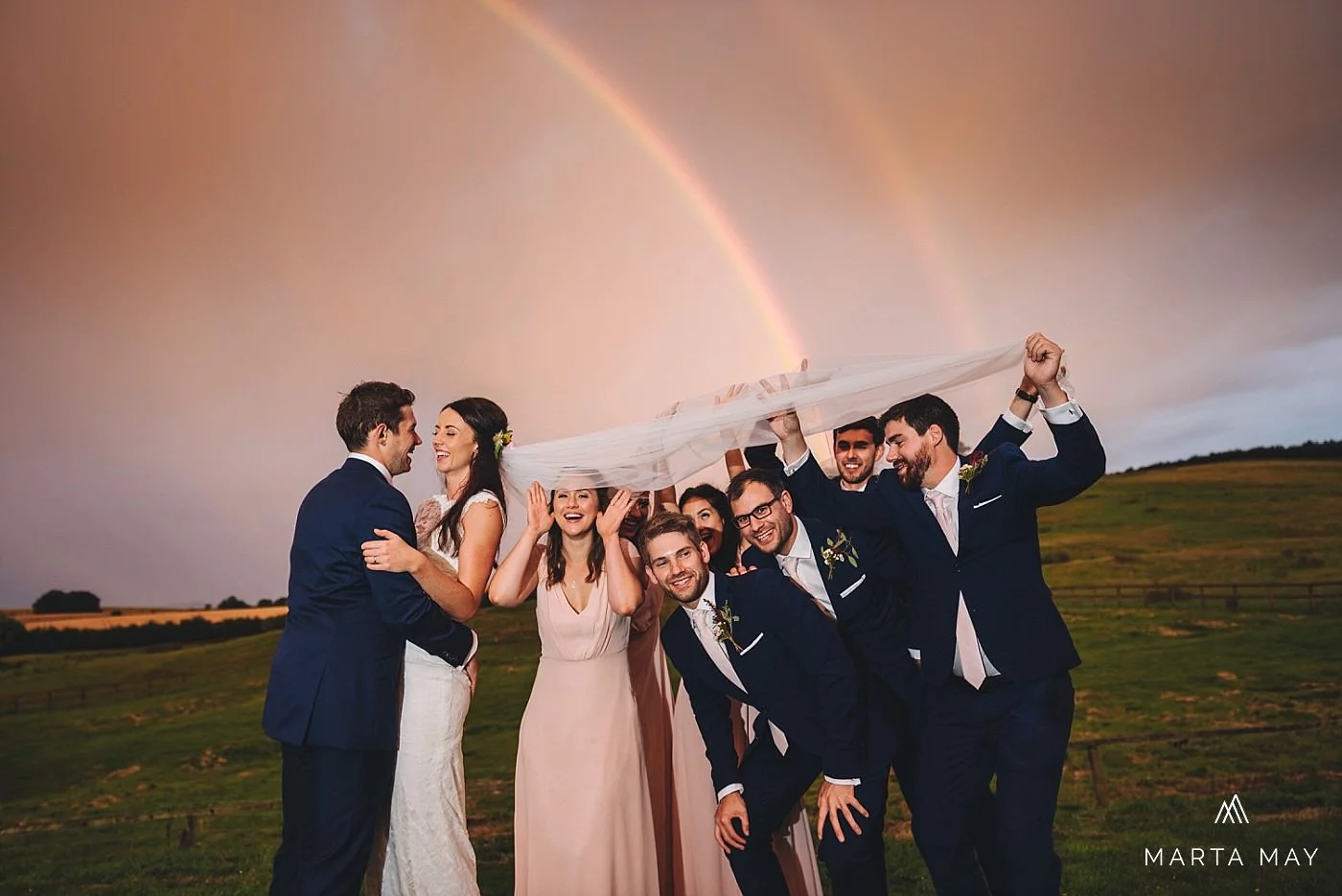 just married bride and groom and their bridesmaids and groomsmen laughing and standing under the veil under the double rainbow