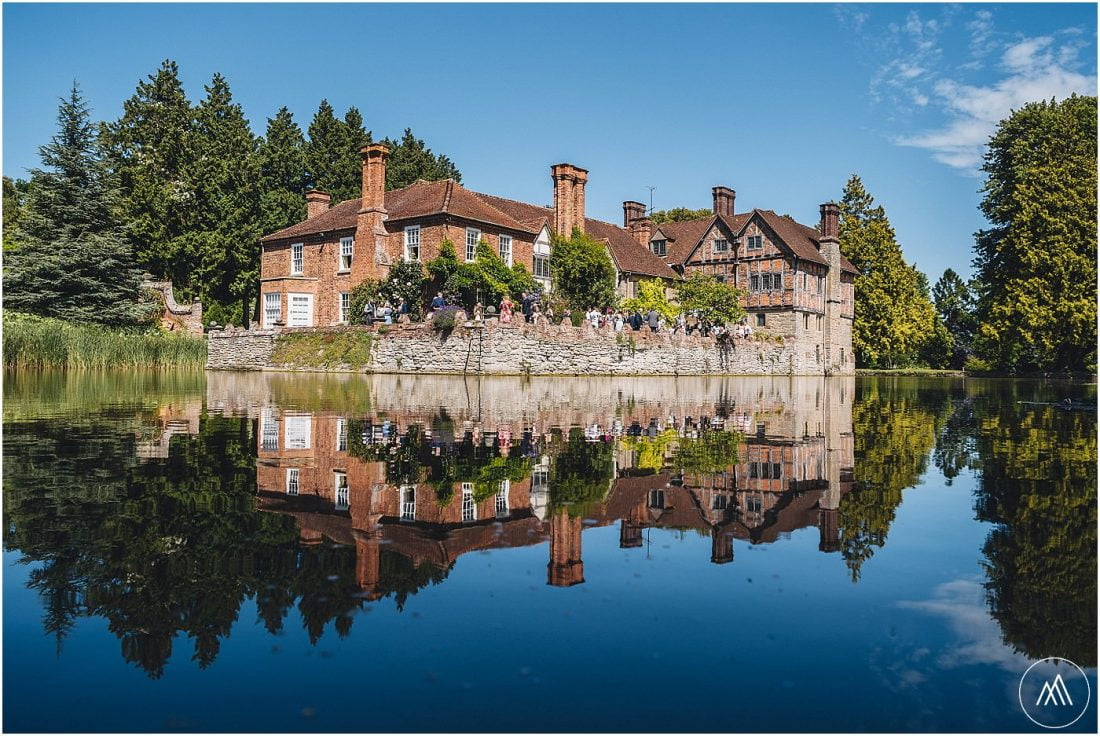 Birtsmorton Court wedding guests enjoying reception drinks on the terrace by the moat in the summer sun. Picture of the house and the sky reflected in the moat