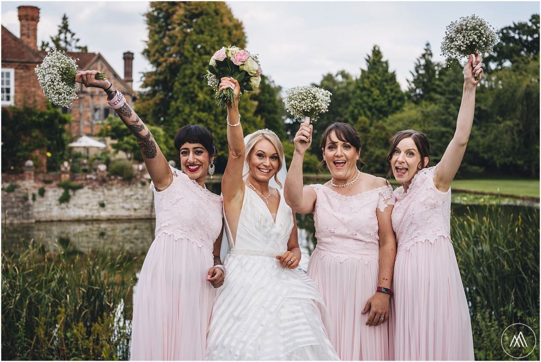 bride and bridesmaids holding flowers up in the air in a group picture in front of the moat at Birtsmorton Court