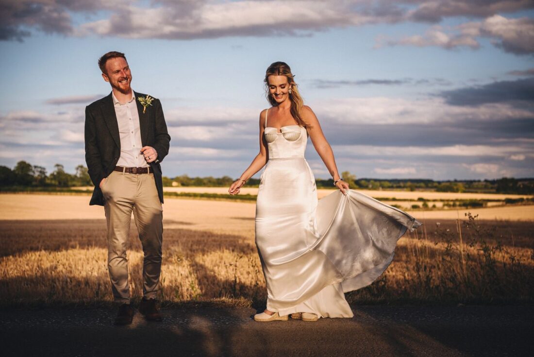 Cripps Barn wedding photography couple standing on the path, bride holding her dress, groom laughing, yellow corn field and blue skies in the background