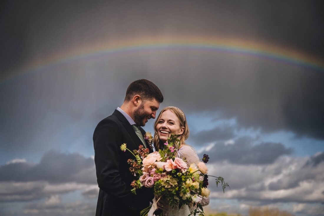 bride and groom hugging and smiling with rain and rainbow in the background