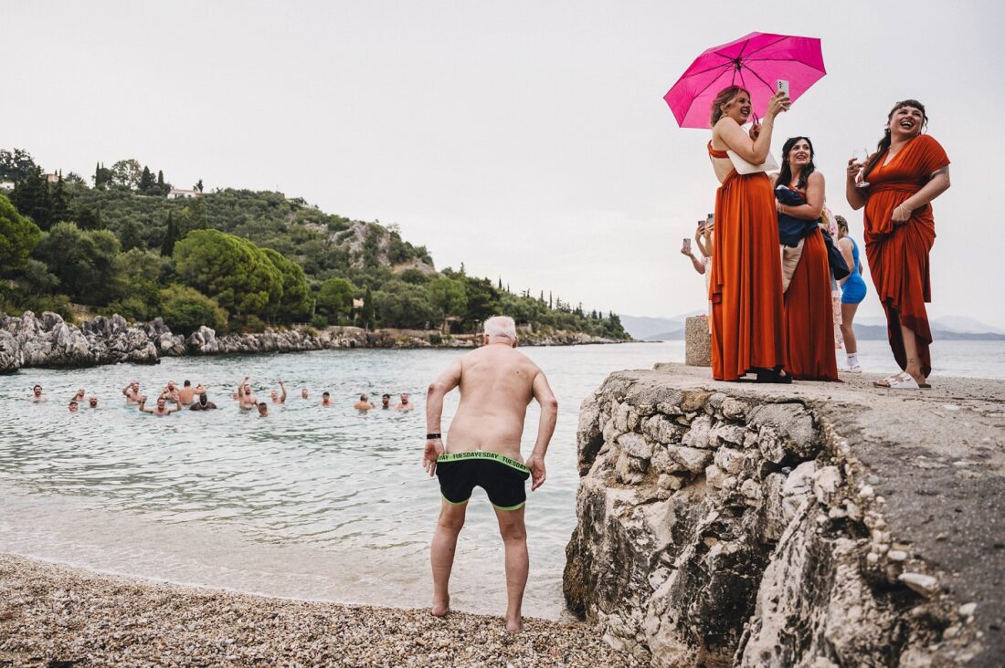 rainy wedding day in Greece, bridesmaids laughing and holding unbrellas, groomsmen swimming and a wedding guest just getting into the water