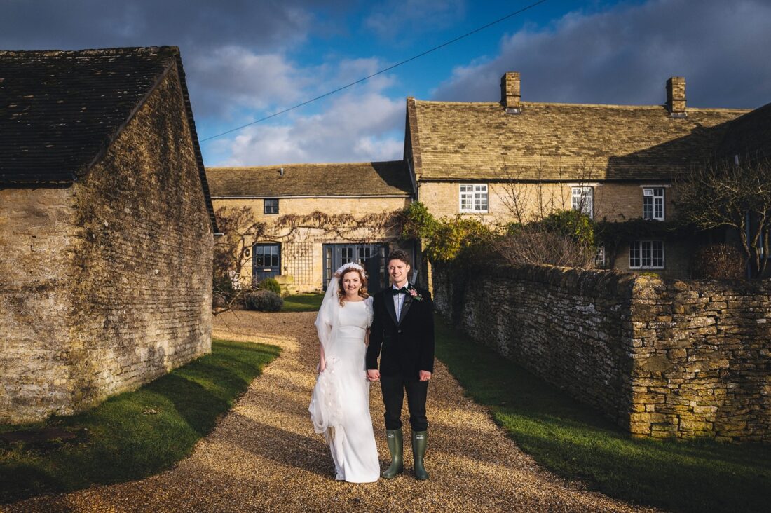 bride and groom standing in front of a Cotswolds barn at Merriscourt, blue skies above them